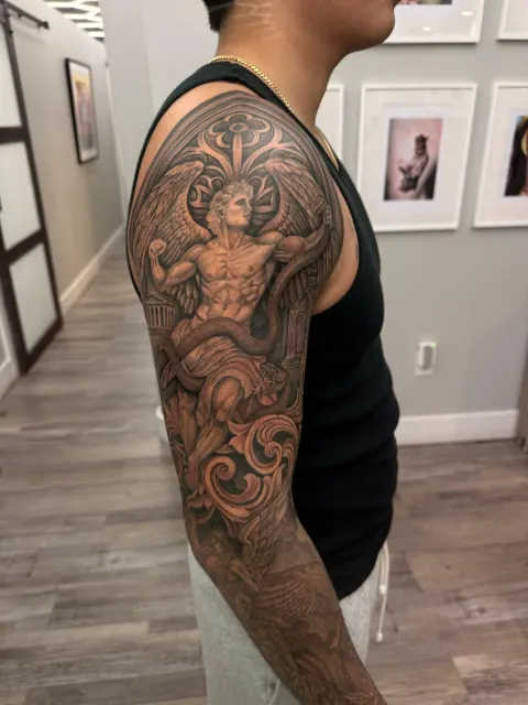 Black and grey upper-arm tattoo of a winged figure inside a baroque filigree frame