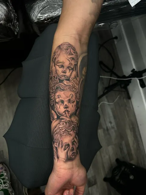 Black and grey forearm tattoo of cherub faces among soft clouds