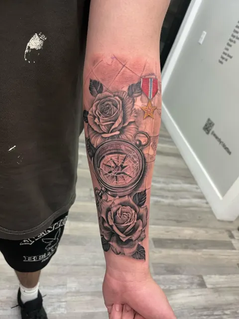 Black and grey forearm tattoo of a compass among roses with a service medal