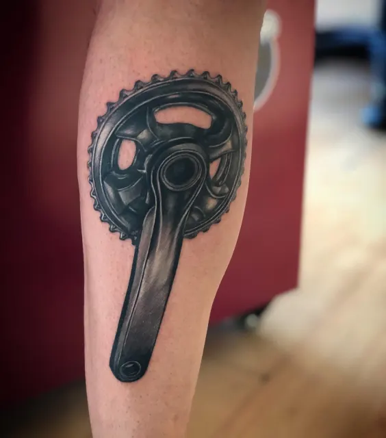 Black and grey forearm tattoo of a bicycle crankset rendered with photographic depth
