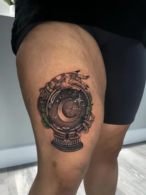 Black and grey thigh tattoo of a crystal ball holding a moonlit scene, with a ship above