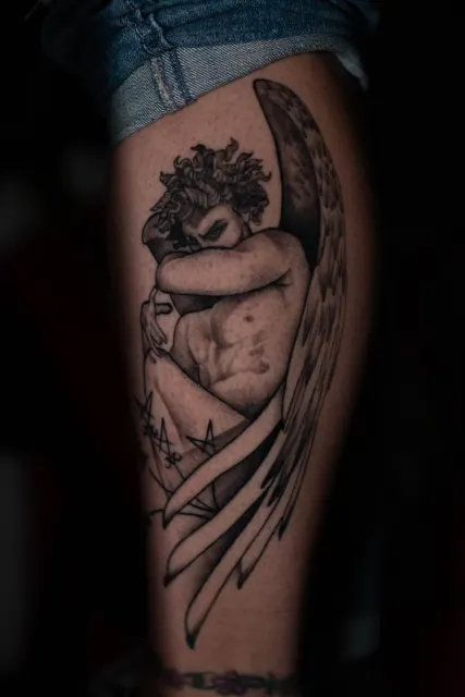 Black and grey thigh tattoo of a winged figure crouched with its head bowed