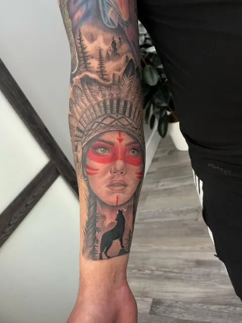 Black and grey forearm portrait of a woman in a feathered headdress with red face paint, beside a howling wolf