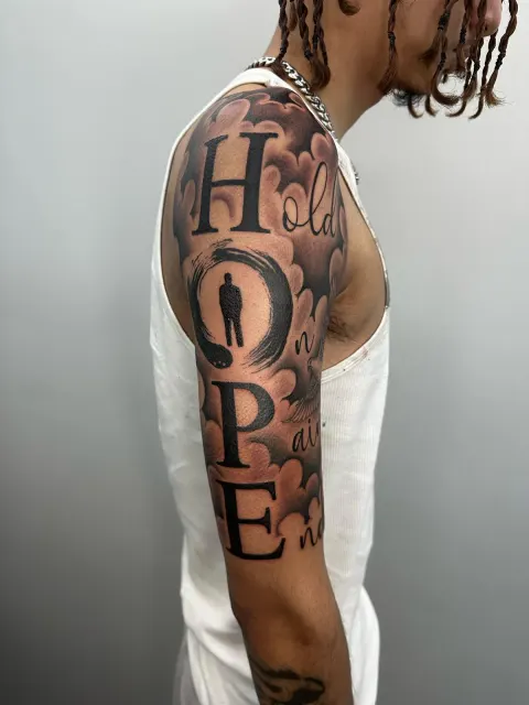 Black and grey upper-arm lettering piece spelling HOPE from the words Hold On, Pain Ends, set in clouds