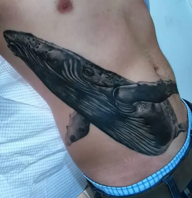 Black and grey rib tattoo of a humpback whale gliding across the side