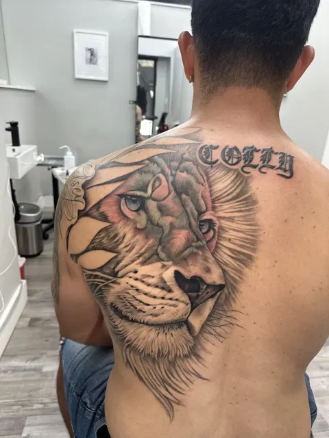 Black and grey upper-back tattoo of a lion beside script lettering