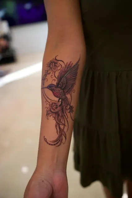 Fine-line forearm tattoo of a phoenix in flight with trailing tail feathers