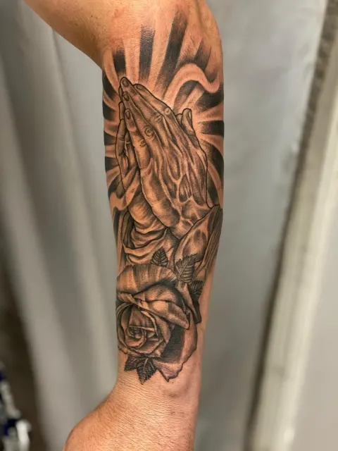 Black and grey forearm tattoo of praying hands over a rose, lit by rays