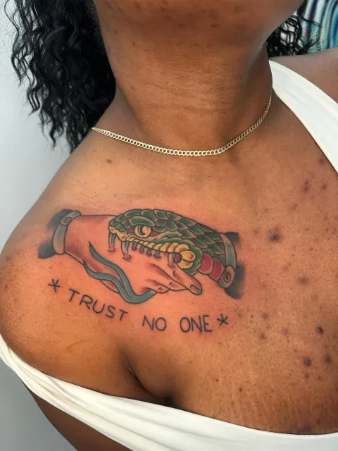 Black and grey collarbone tattoo of a serpent head above lettering reading Trust No One