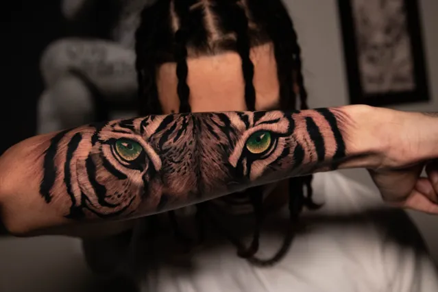 Black-and-grey forearm tattoo centered on tiger eyes with green-gold irises and bold black stripes