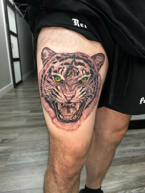 Black and grey thigh tattoo of a roaring tiger with green eyes