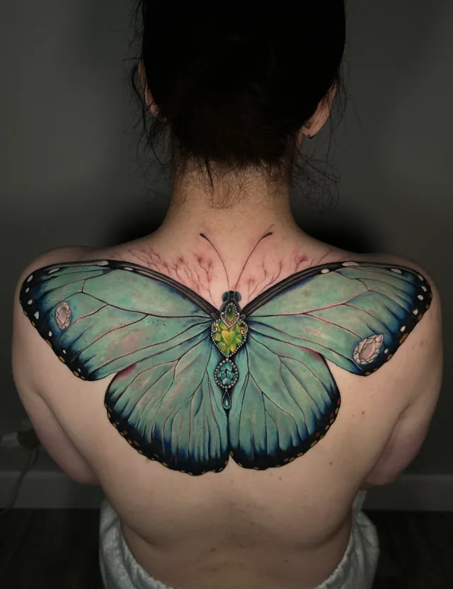 Large turquoise butterfly tattoo across the upper back with blue-edged wings and green and blue jewel-like accents