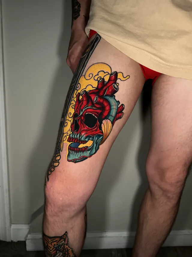 Color thigh tattoo of an anatomical red heart above a blue-toned skull