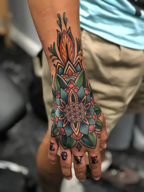 Ornamental color mandala across the back of the hand in teal and red, with lettering across the knuckles