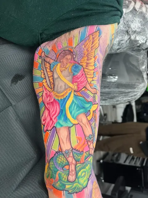 Color upper-arm tattoo of a winged archangel over a fallen figure, rendered in saturated rainbow tones