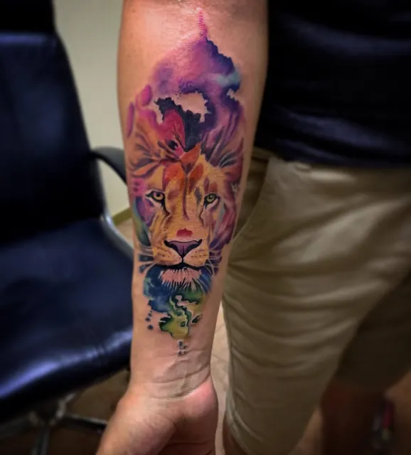 Forearm tattoo of a lion profile dissolving into watercolor washes of violet and teal