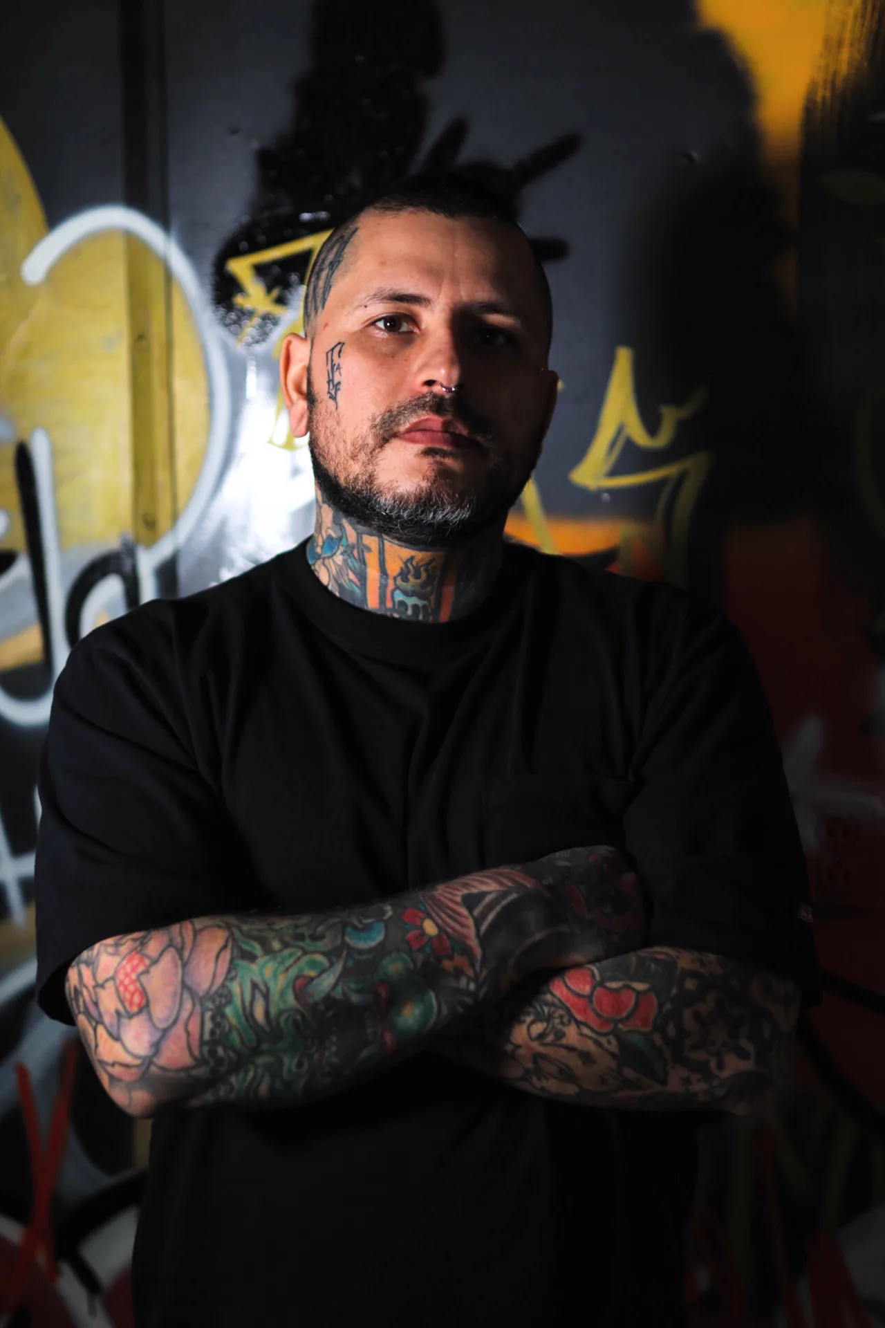 Portrait of Dymon Jimenez in a black shirt with tattooed arms crossed, in front of a black-and-yellow graffiti wall