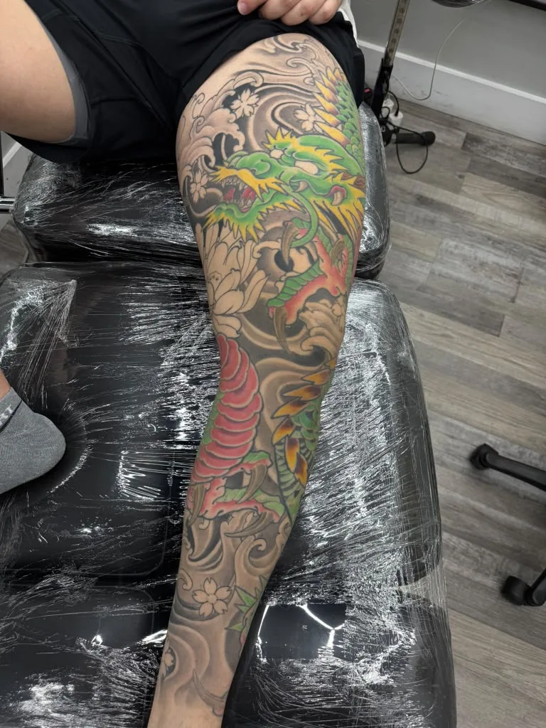 Client on the studio bench during a full-leg Japanese dragon session, protective film over the furniture