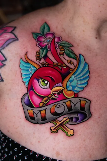 Traditional chest tattoo of a pink swallow over a banner reading Mom, with a cross below