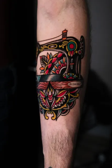 Traditional forearm tattoo of a black sewing machine with red flowers and a lotus below